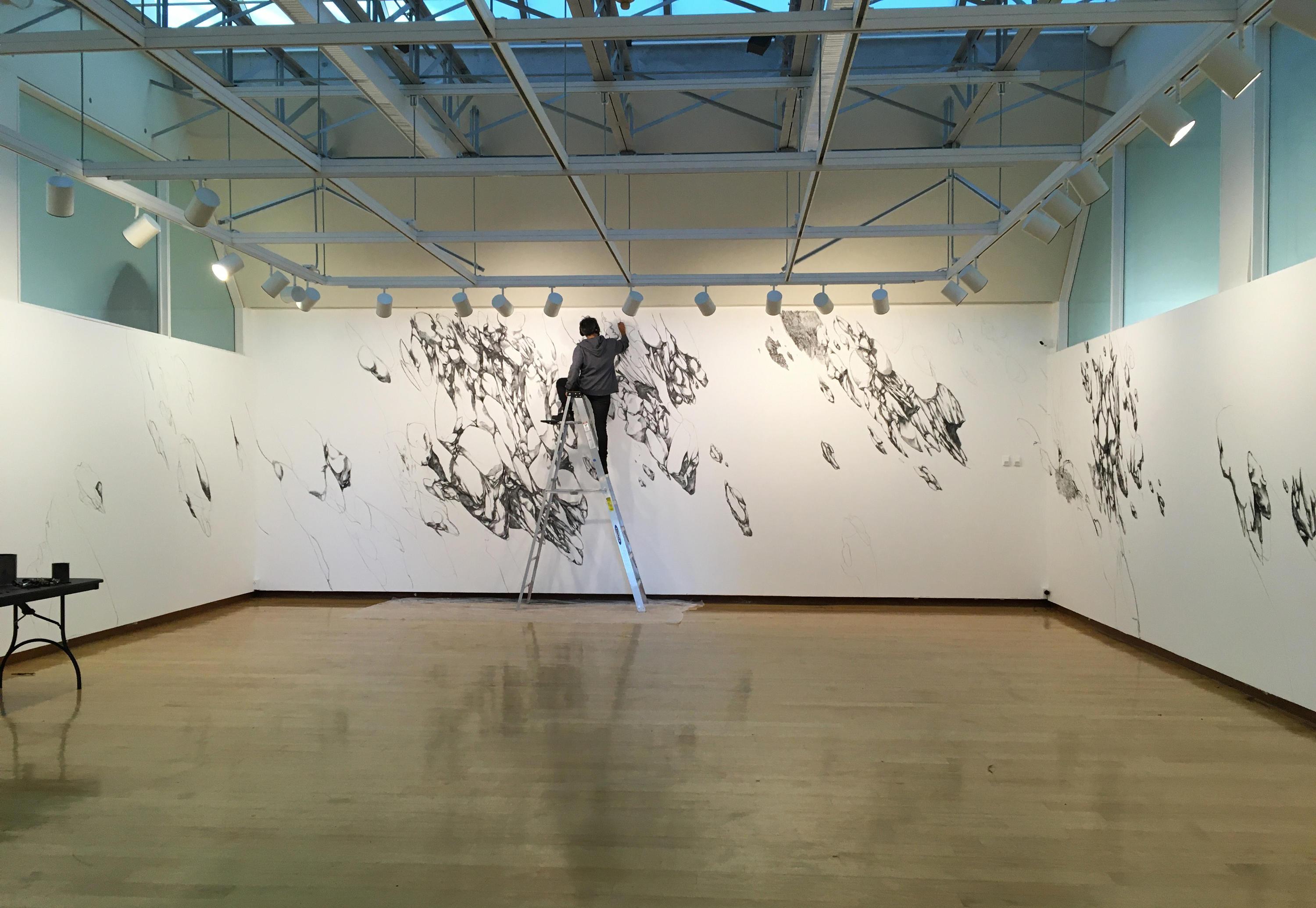 person in gray and black clothing standing at the top of a ladder, 回到镜头前, drawing on a wall that already has charcoal rocklike drawings on it