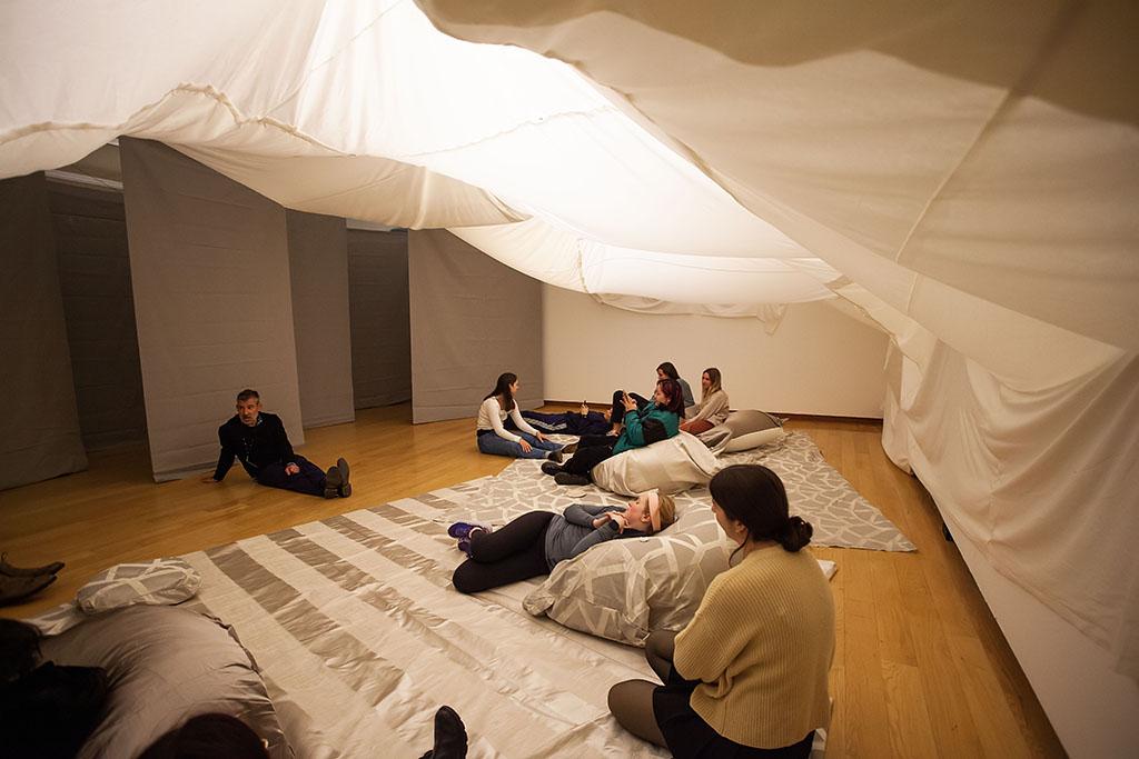 戏剧性地垂下白色织物天花板, 从后面点燃, above a group of people sitting on a floor strewn with gray and white fabric and cushions; behind them is a series of gray hanging fabric panels