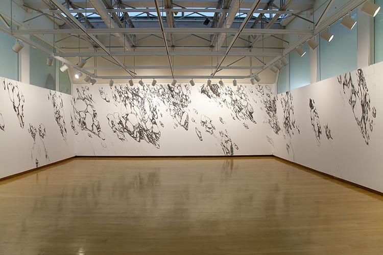 一个白, wood-floored gallery with enormous charcoal drawing of rocks flying through air on the walls