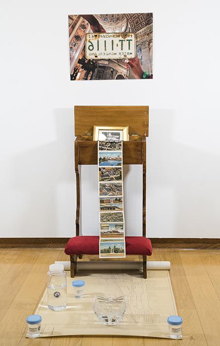 photo of two hands in a chapel surrounding a Michigan 'Water Wonderland' license plate mounted on the wall; below, 一张填满蜉蝣的祈祷凳，地板上放着一张地图，上面放着各种盛水的容器, 包括样品罐