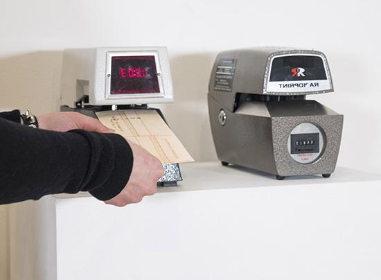 two old fashioned punch clock machines on a pedestal; hands insert a punch card into the machine on the right